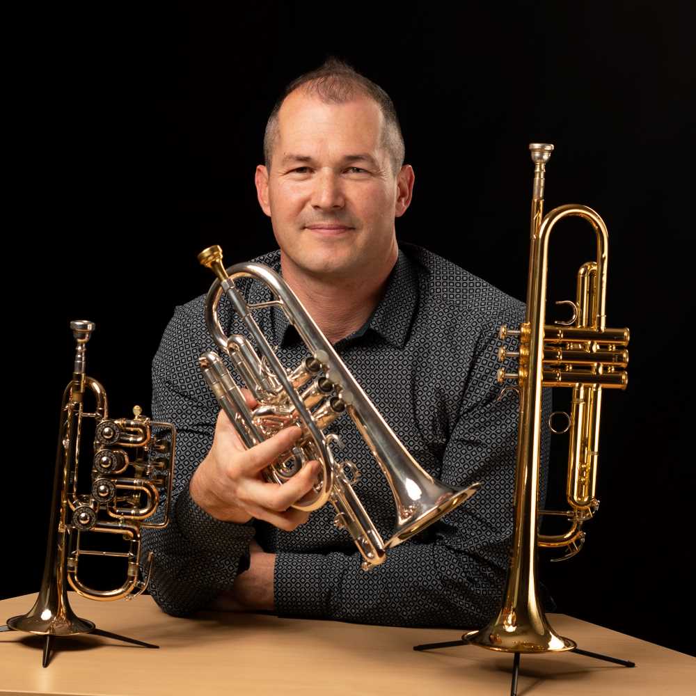 Laurent Jammes attablé avec trois cuivres : cornet, trompette et trompette piccolo. Toulouse, 2025.